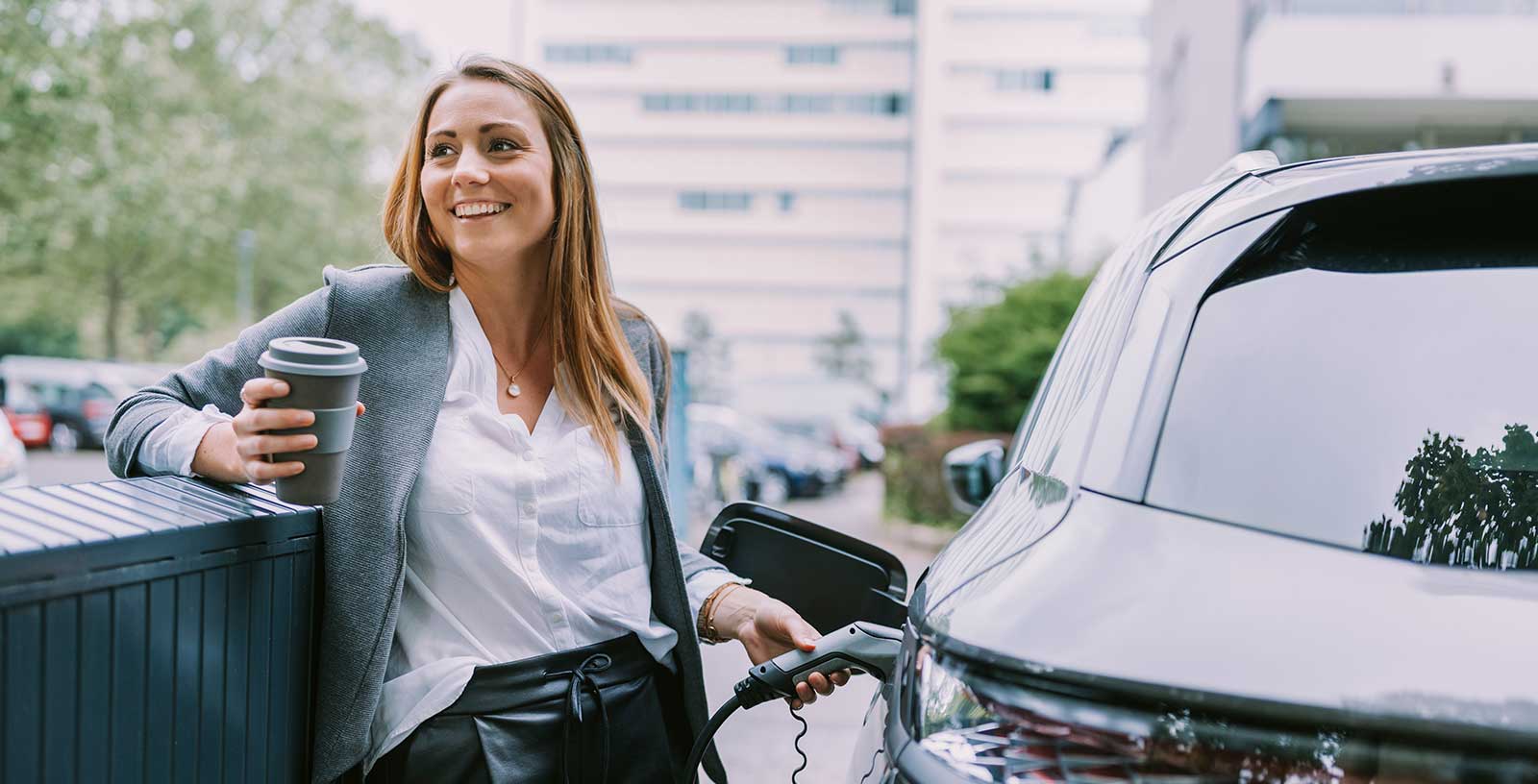 Eine Frau mit Mehrweg-Kaffeebecher in der Hand lädt ihr Elektroauto