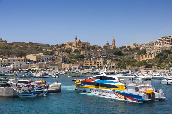 Gozo Ferry Port with ferry and boats in harbour, azure blue sea and Maltese buildings behind